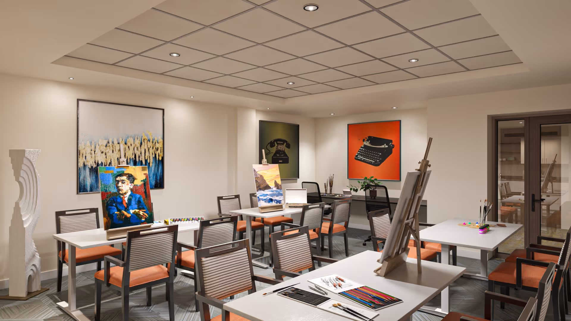 A well-lit art room with multiple tables and chairs arranged for painting activities. Several easels hold colorful paintings, and art supplies like colored pencils and brushes are placed on the tables. The walls are decorated with framed artwork, including images of a vintage telephone and a typewriter. The room has a modern ceiling with recessed lighting and a glass door leading to another room.
