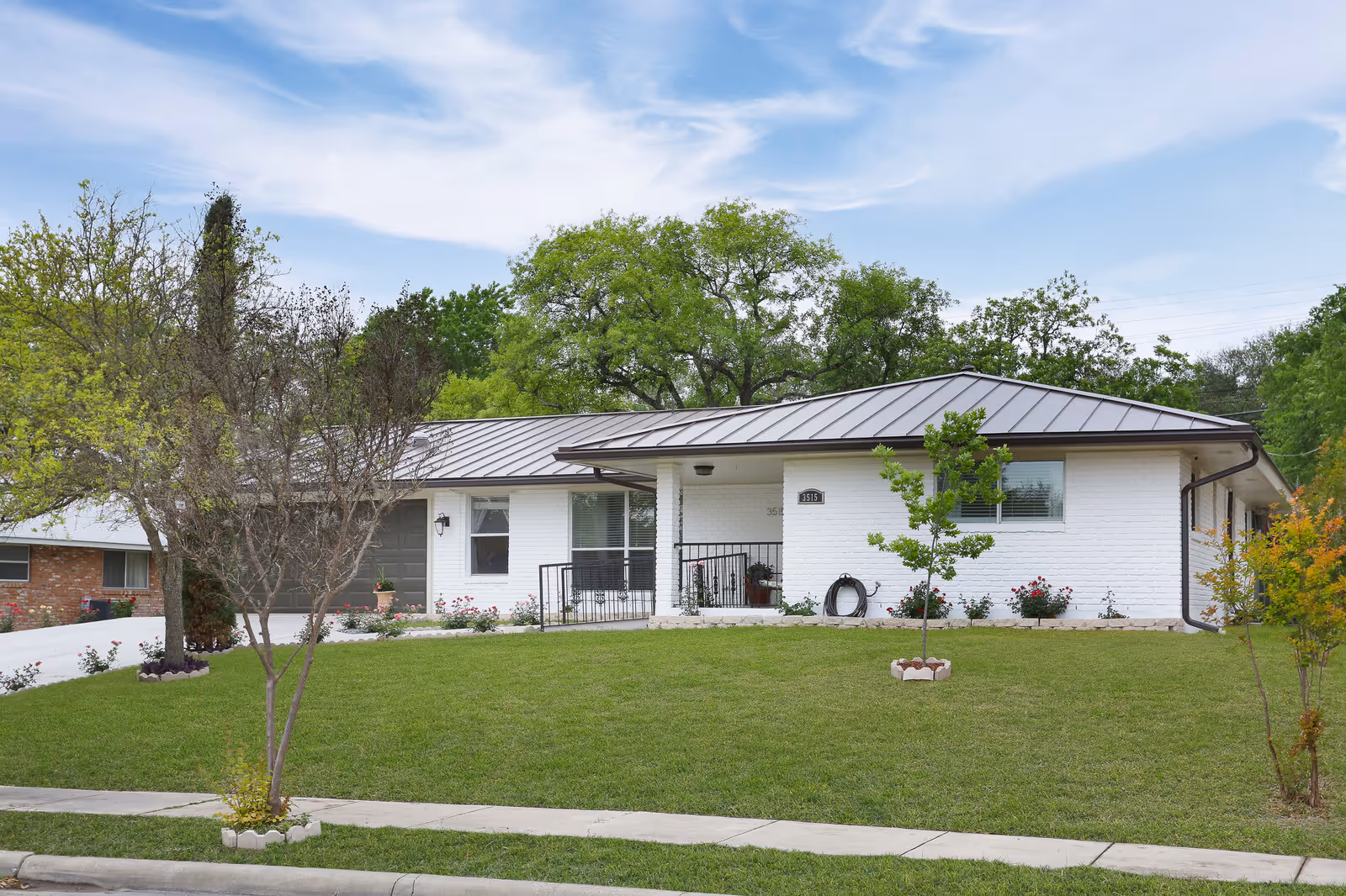 Single-story white brick ranch-style house with a metal roof, front porch, lawn, and trees.