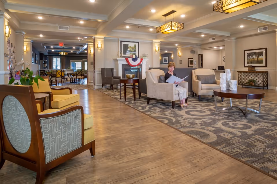 A spacious and well-lit senior living facility common area with comfortable armchairs and a patterned carpet. An elderly woman is seated in one of the armchairs reading a book. The room features warm lighting fixtures, framed artwork on the walls, and a fireplace decorated with a patriotic banner. In the background, there is a dining area with tables and chairs.