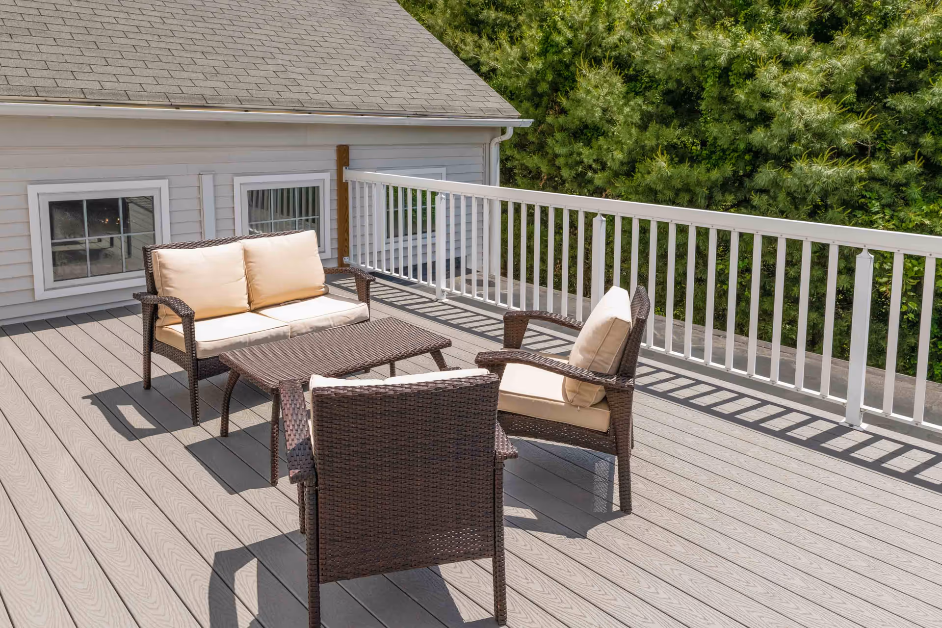 Outdoor deck with wicker loveseat, chairs, and a coffee table beside a white railing and trees.