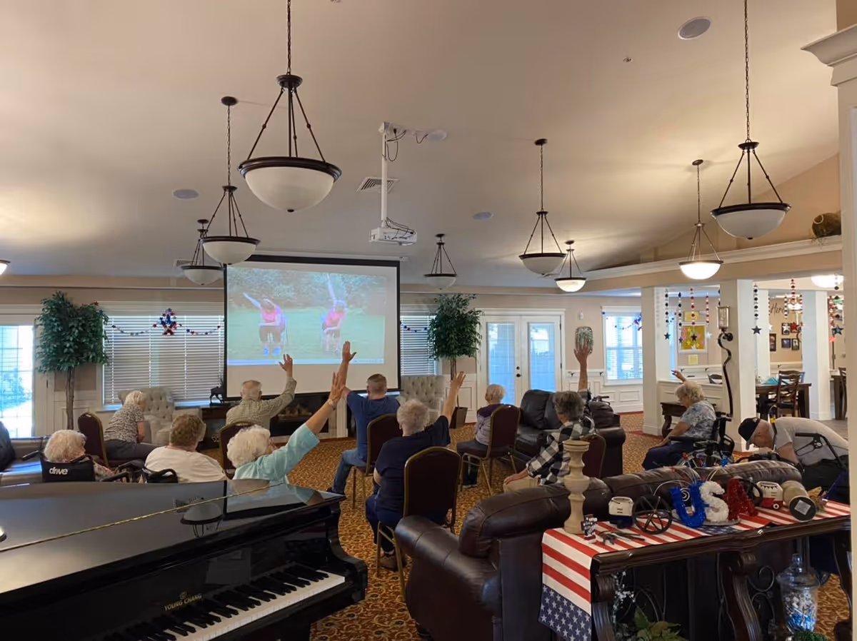 A group of elderly people seated in chairs and wheelchairs in a common room participating in a seated exercise session, following along with an exercise video projected on a large screen. The room is well-lit with hanging light fixtures, decorated with plants, and has comfortable seating including leather sofas and armchairs.