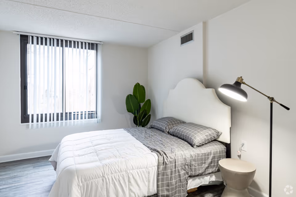 Bright minimalist bedroom with a bed, gray bedding, a tall floor lamp, a potted plant, and a window with vertical blinds.