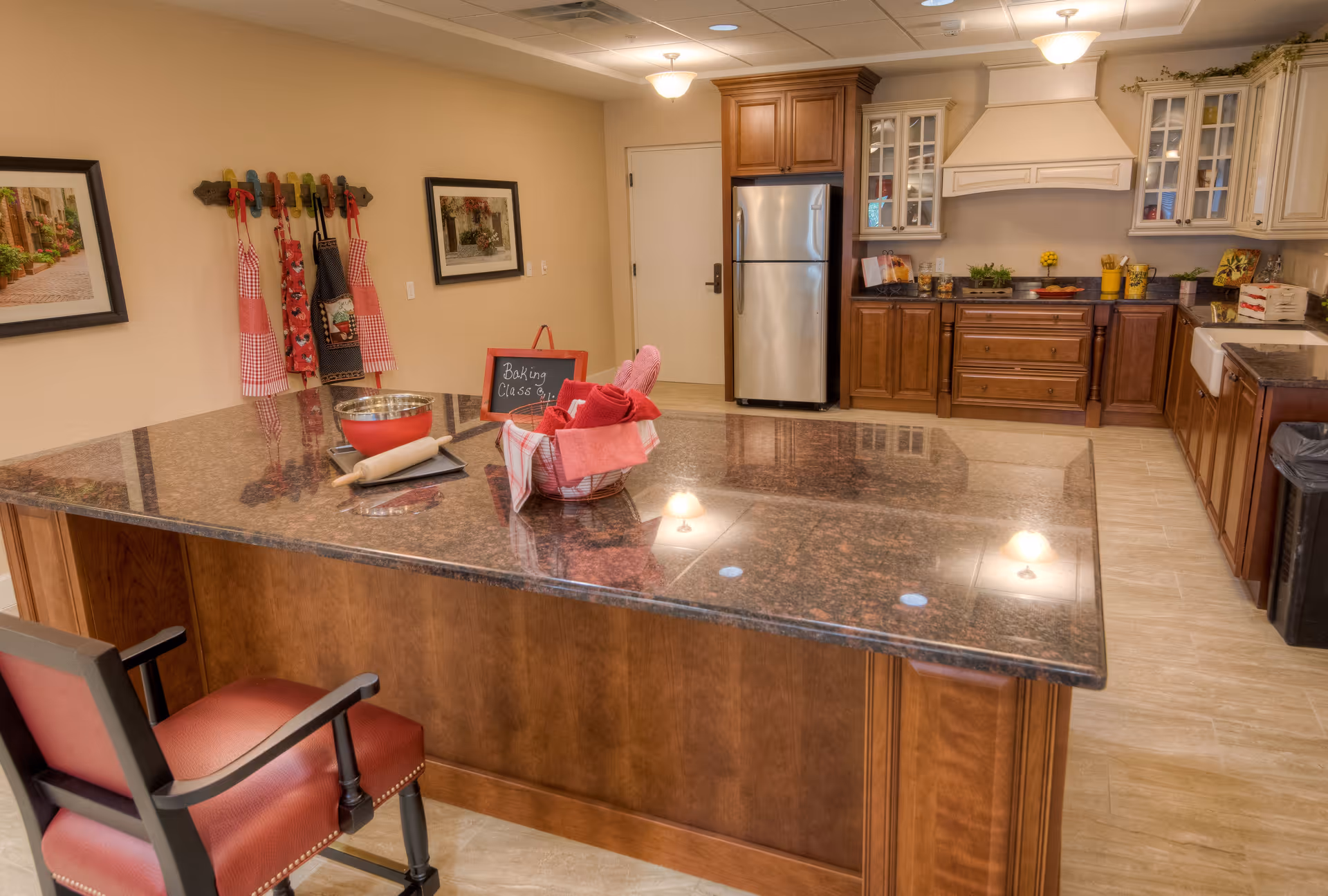 A spacious kitchen with a large granite island countertop in the center. The island has a red chair, a rolling pin, a mixing bowl, and a basket with red towels. On the wall, there are colorful aprons hanging and two framed pictures. The kitchen features wooden cabinets, a stainless steel refrigerator, and a stove with a white range hood. The floor is tiled, and the room is warmly lit with ceiling lights.