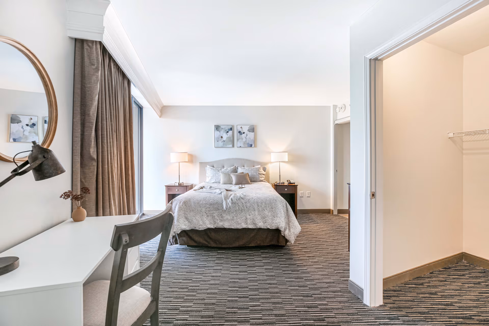 Bright, modern bedroom with a bed centered between nightstands and lamps, a desk and chair in the foreground, and an open closet to the right.