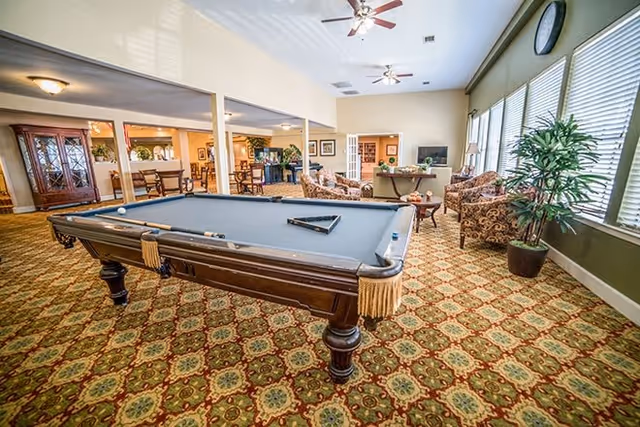 A spacious communal lounge featuring a pool table, patterned carpet, seating areas, and large windows.