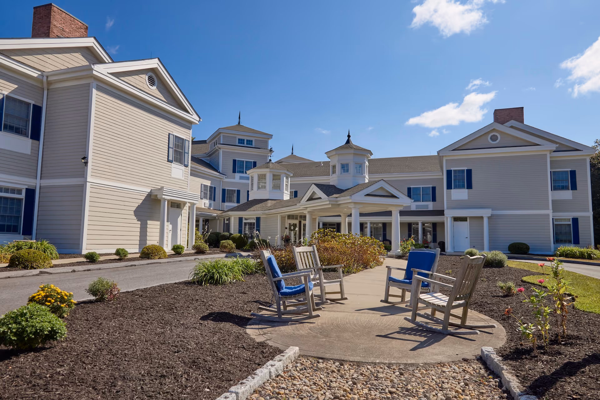 Exterior view of Monarch Southbury facility on a sunny day with a clear blue sky. The building is light-colored with multiple windows and architectural details including small towers and a covered entrance. In the foreground, there is a circular concrete area with four wooden rocking chairs, two of which have blue cushions, surrounded by landscaped garden beds with mulch and plants.