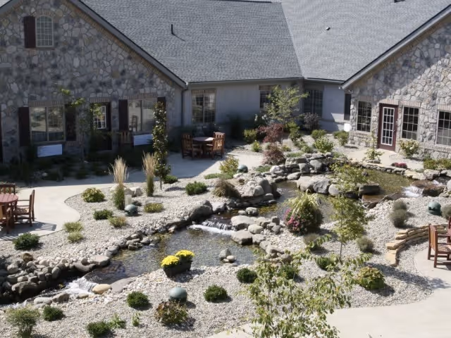 Outdoor courtyard area at The Inn at CoalRidge featuring a stone building with multiple windows and doors, a landscaped garden with rocks, small plants, and a flowing water feature, along with several wooden tables and chairs placed on paved walkways.