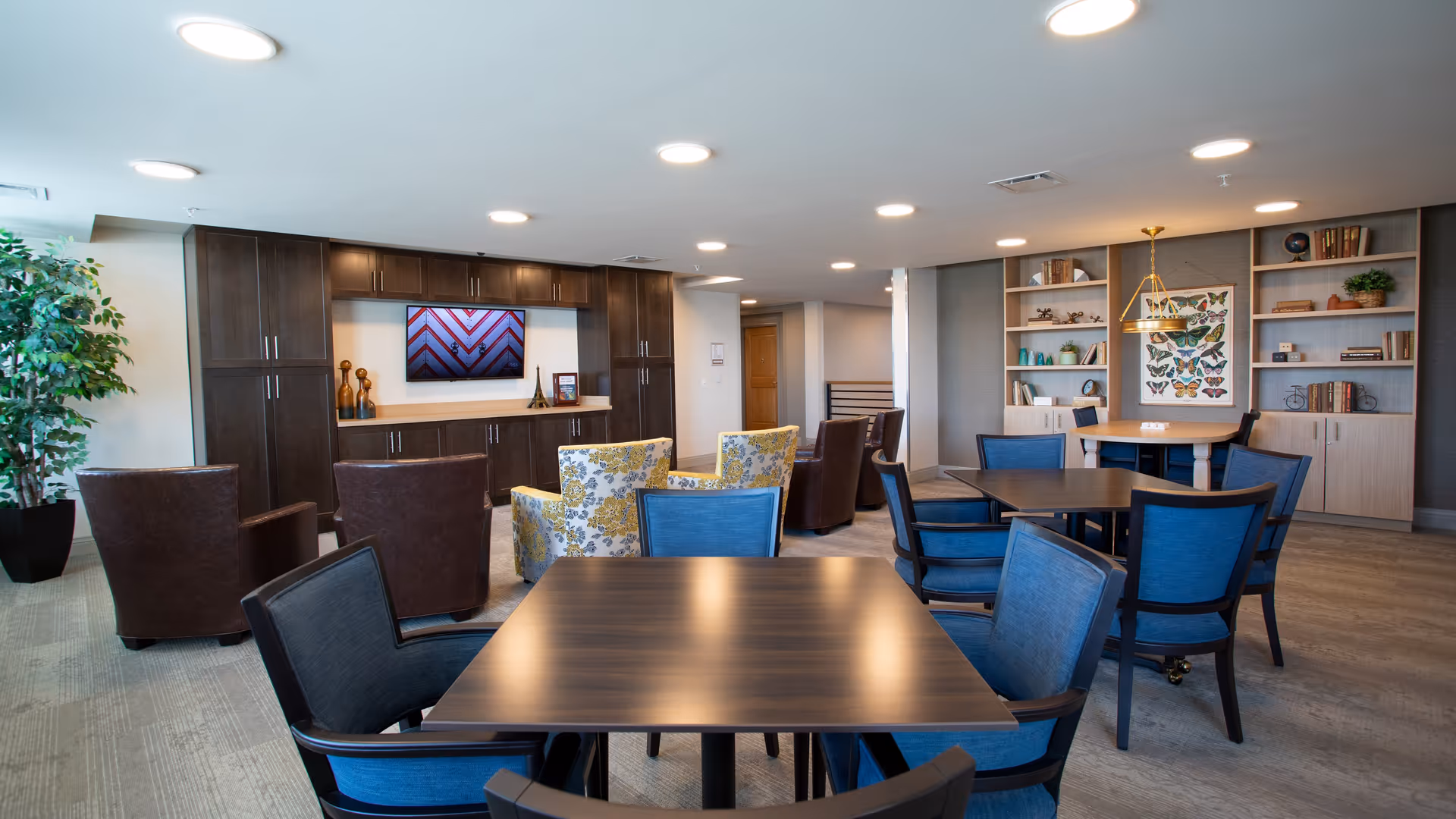 Bright communal lounge with multiple tables and blue chairs, armchairs, a wall-mounted TV and built-in shelving.