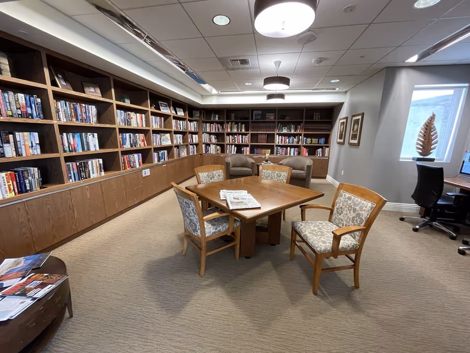 A cozy library room with wooden bookshelves filled with books along the walls. In the center, there is a wooden table with four cushioned chairs around it, and newspapers placed on the table. Two armchairs and a small table with a lamp are positioned against the back wall. The room has carpeted flooring, framed pictures on the wall, a window with a decorative sculpture on the sill, and a computer workstation with an office chair near the window. Ceiling lights illuminate the space.