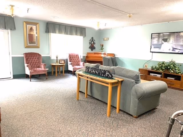 A cozy living room area in Cambridge Court Assisted Living featuring a gray couch with a wooden table behind it displaying a decorative 'BLESSINGS' sign. Two pink upholstered armchairs are positioned near a window with green valances. A wooden piano is against the far wall, and a flat-screen TV is mounted on the wall above a wooden media console with plants and decor. The room has light green walls with white trim and a patterned carpet.