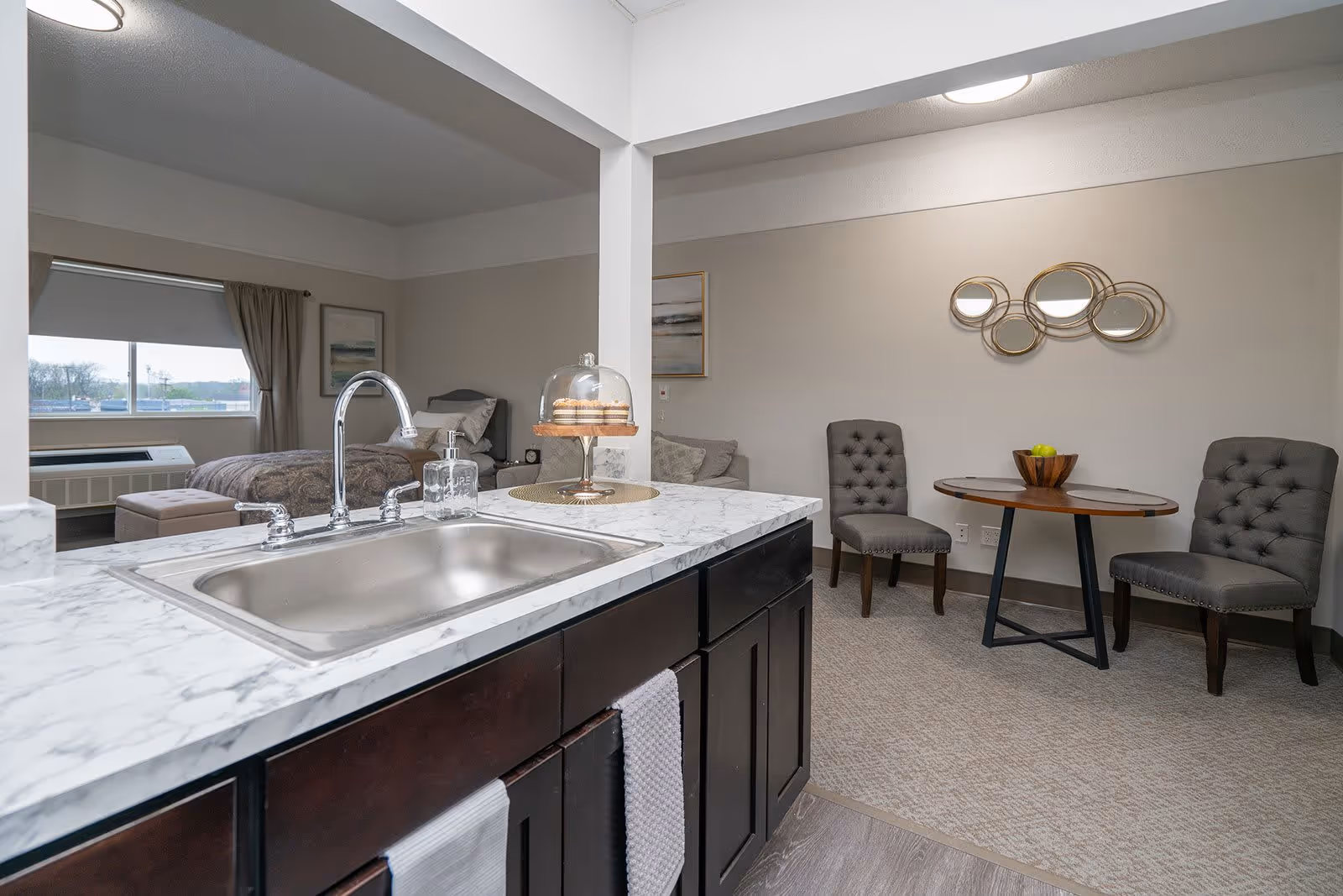 Interior view of a senior living facility apartment showing a kitchen sink with marble countertop in the foreground, a small round dining table with two gray upholstered chairs, and a bedroom area with a bed and window in the background.