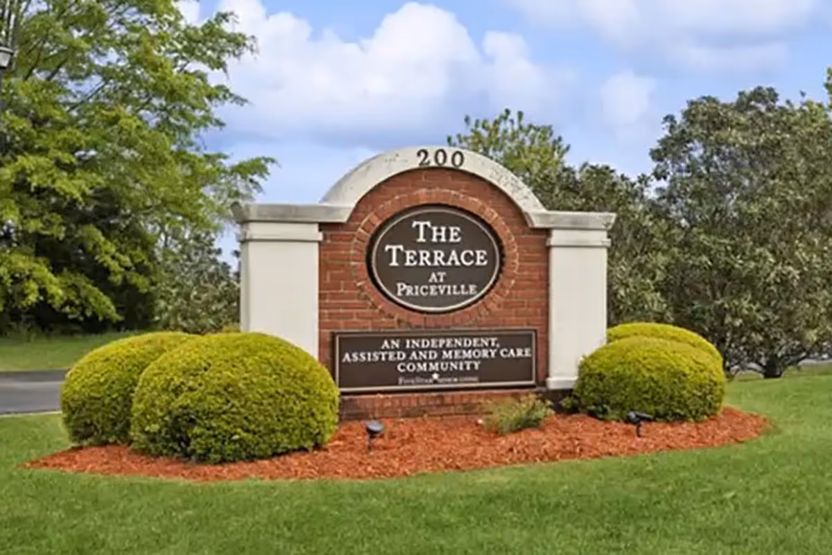 A brick and stone sign for The Terrace at Priceville, an independent, assisted, and memory care community, surrounded by neatly trimmed bushes and green grass under a partly cloudy sky.