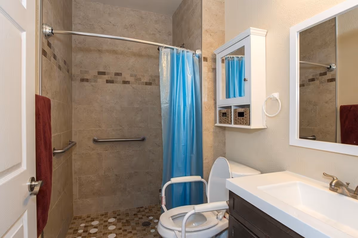 A bathroom with beige tiled walls and floor, featuring a walk-in shower with a blue shower curtain and grab bars. There is a white toilet with arm supports, a white vanity with a sink and faucet, a wall-mounted cabinet with two wicker baskets, a large mirror, and a red towel hanging on the wall.