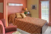 A cozy bedroom with a patterned bed, decorative pillows, a bedside table with lamp and photos, a striped armchair, and a window with blinds.