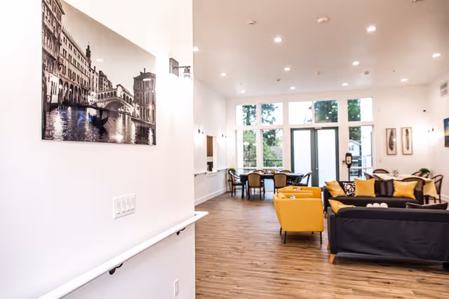 Bright and spacious senior living common area with wooden flooring, large windows letting in natural light, yellow and black sofas, a dining table with chairs, and a black and white photograph of a canal scene on the wall.