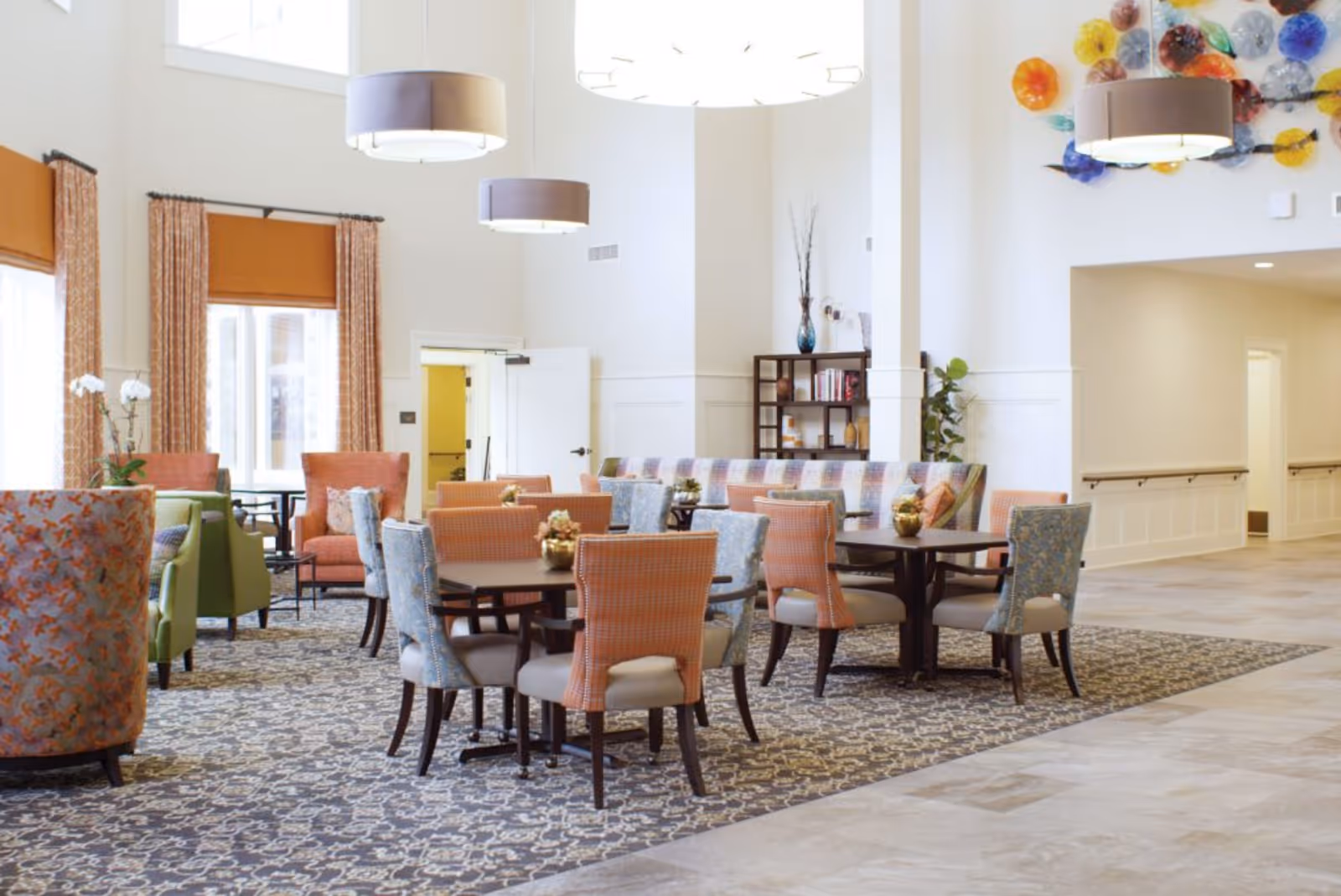 A bright and spacious common area with multiple round tables surrounded by colorful upholstered chairs in shades of orange, blue, and green. The room features large windows with orange curtains, modern pendant lights hanging from the high ceiling, a patterned carpet, and a decorative wall installation with colorful glass pieces. There is also a bookshelf and some plants in the background.