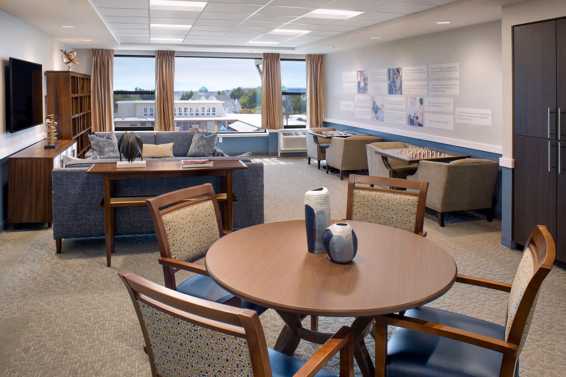 Communal senior living lounge with a round table and chairs in the foreground, sofas and game tables near large windows overlooking the neighborhood.