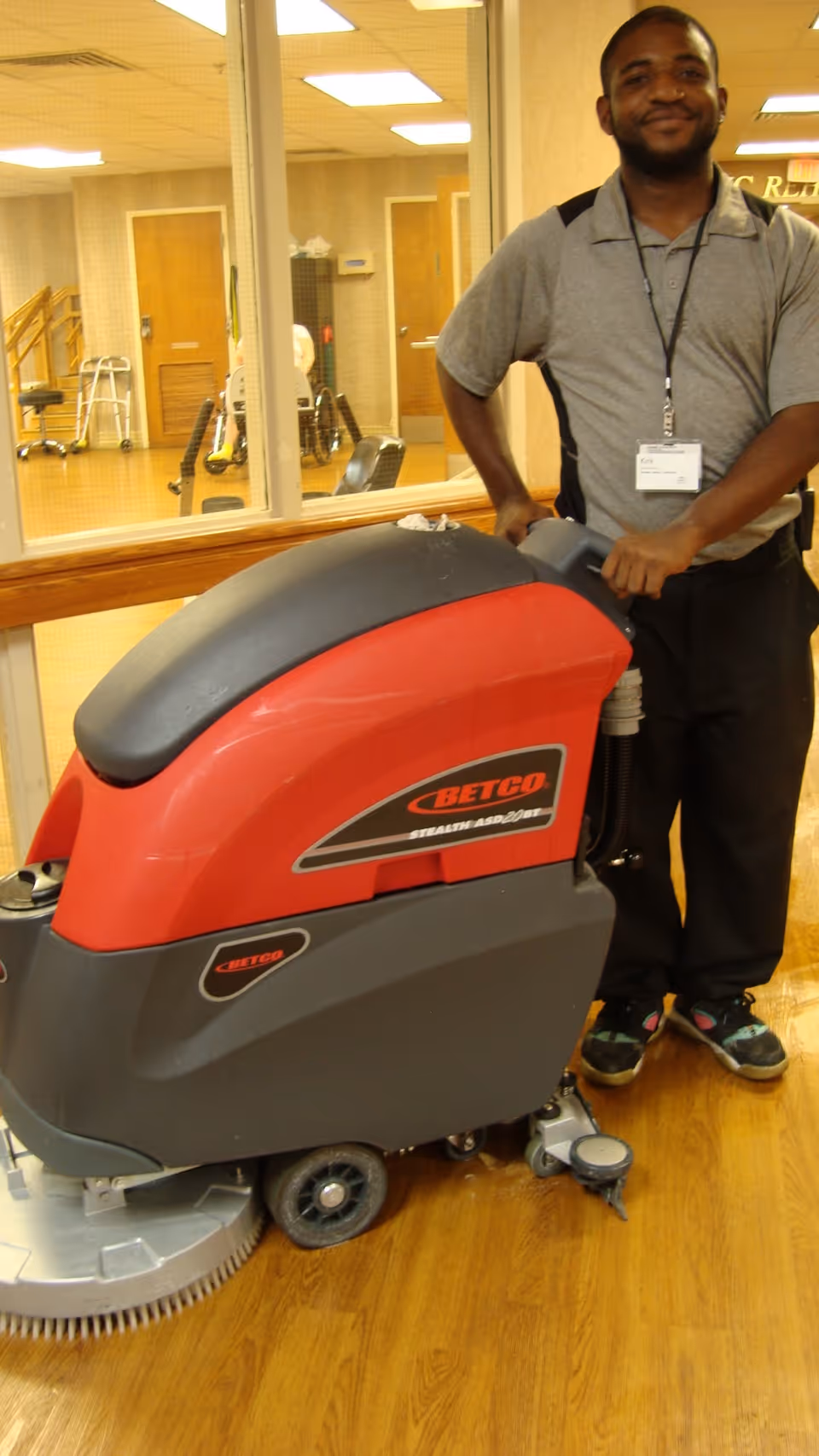A man standing indoors next to a large red and gray floor cleaning machine labeled BETCO. The man is smiling and wearing a gray and black shirt with a name badge. The background shows a room with wooden floors, glass windows, and some medical equipment like walkers and wheelchairs.