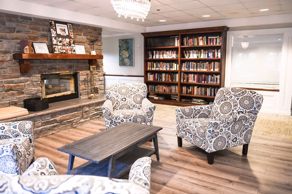 A cozy living room area with four patterned armchairs arranged around a wooden coffee table. There is a stone fireplace with a wooden mantel displaying framed photos and certificates. A large bookshelf filled with books is against the wall, and a chandelier hangs from the ceiling.