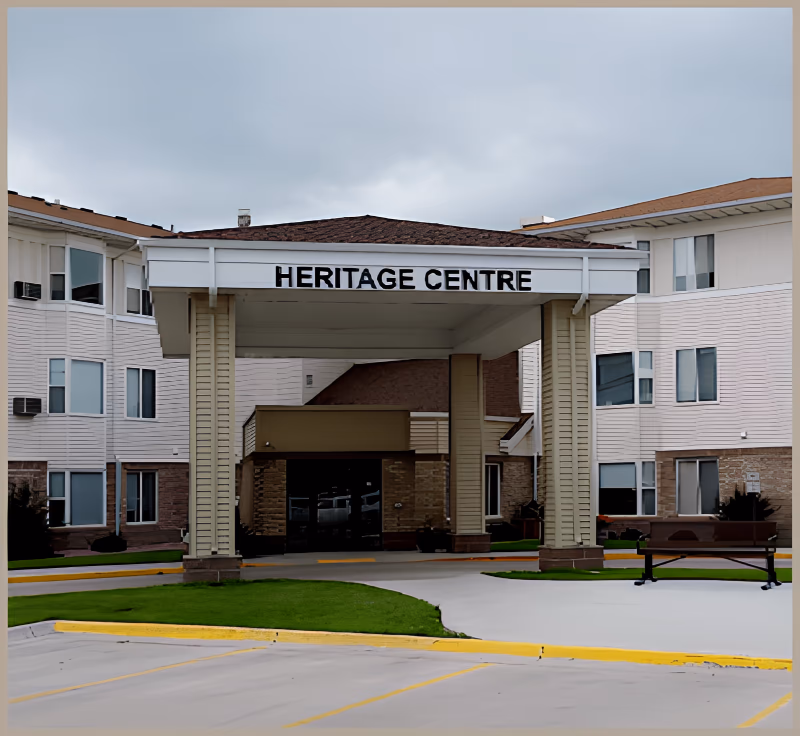 Front entrance of the Heritage Centre building with a covered drop-off area, surrounded by a multi-story structure with windows and a bench on the right side near the entrance.