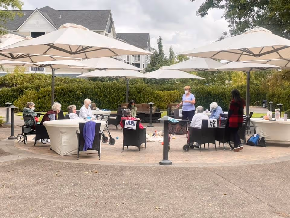A group of elderly people sitting in a circle outdoors under large white umbrellas. Some are seated in chairs and others in wheelchairs, with two caregivers standing nearby. The setting is a paved patio area with greenery and residential buildings in the background.