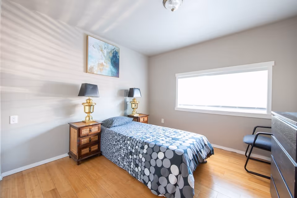 A bright bedroom with a single bed covered in a blue and gray polka dot patterned bedspread. There are two wooden nightstands on either side of the bed, each with a gold lamp with a black lampshade. A large window with white trim lets in natural light. A modern abstract painting hangs on the wall above the bed. There is a black chair and a dark dresser on the right side of the room. The floor is wooden.