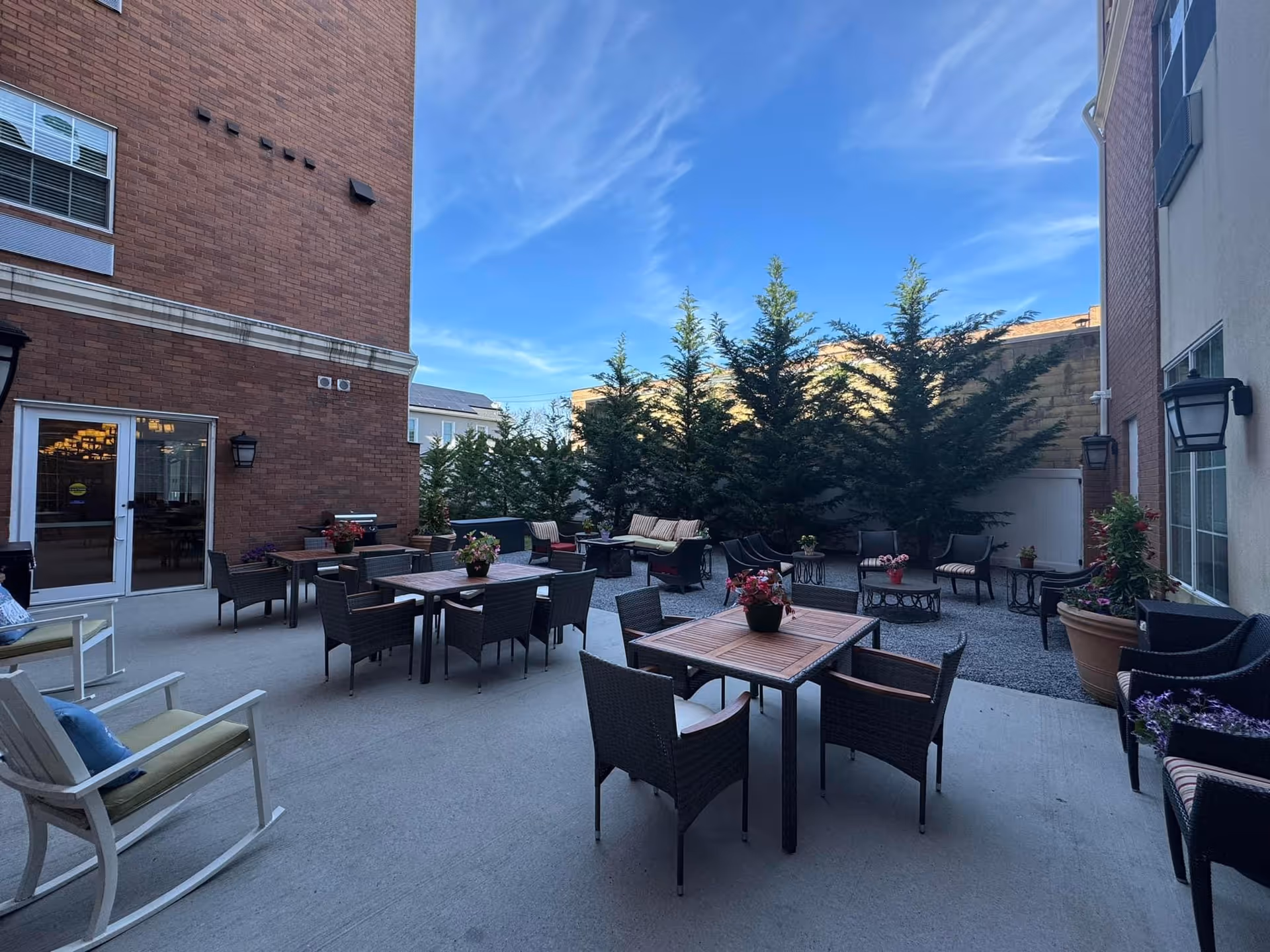 Outdoor patio area at Village Walk Senior Living with multiple tables and chairs arranged for seating. There are potted plants on the tables and around the patio. The space is enclosed by buildings and tall evergreen trees, under a clear blue sky.