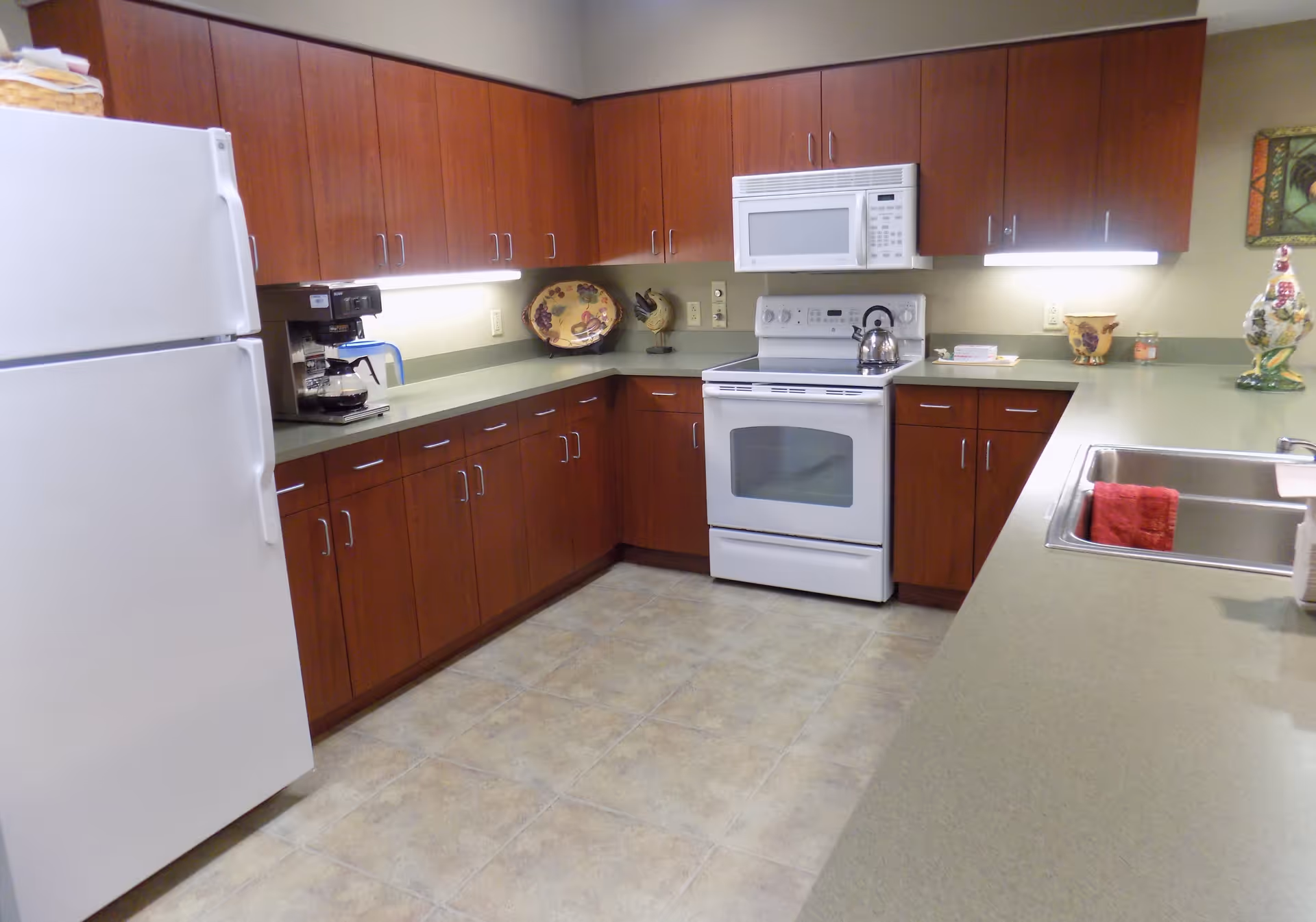 A clean kitchen with wooden cabinets, a white refrigerator, a white stove with a kettle on it, a white microwave above the stove, a coffee maker on the counter, and a double sink with a red towel hanging inside. The countertops are light green, and the floor is tiled. There are decorative items on the counter and a framed picture on the wall.