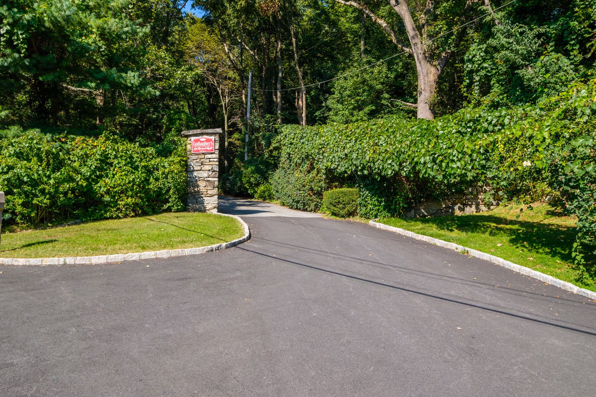 Asphalt driveway entrance bordered by green hedges and trees with a stone pillar sign reading 'Gallagher's'.