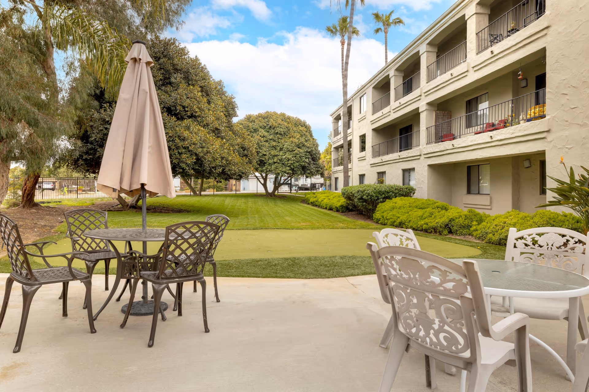 Outdoor patio area at Laguna Estates Senior Living with metal and plastic chairs around tables, one table with a closed beige umbrella. The patio overlooks a well-maintained lawn with trees and shrubs, adjacent to a three-story residential building with balconies.