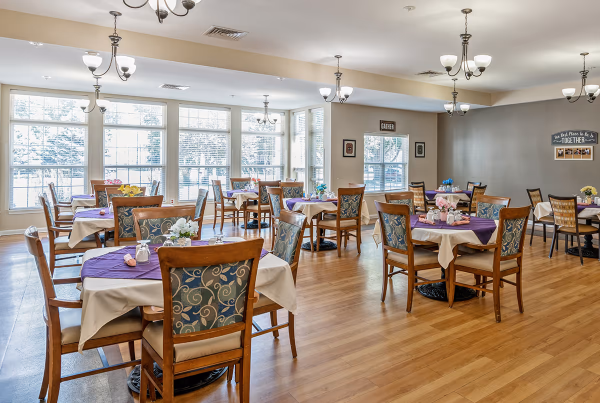 A bright dining room with multiple wooden tables and chairs arranged neatly. Each table is covered with a white tablecloth and a purple overlay, decorated with small flower arrangements and place settings. Large windows allow natural light to fill the room, and several ceiling light fixtures hang overhead. The walls have framed pictures and signs, including one that says 'Gather' and another that reads 'The Best Place to Be is Together.'
