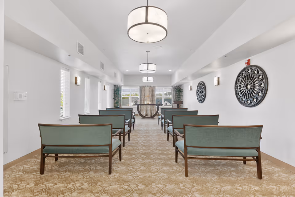 A bright, well-lit room with rows of green cushioned benches facing a small table near large windows with sheer curtains. The room has white walls adorned with decorative circular wall art and modern ceiling lights. There are plants near the windows, and the floor is covered with a patterned beige carpet.