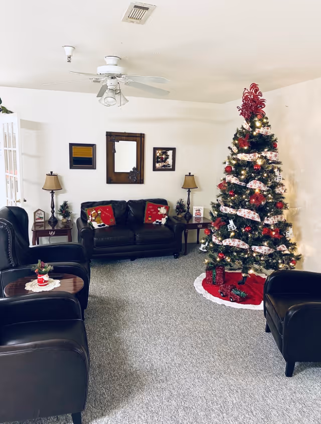 A cozy living room decorated for Christmas with a large Christmas tree adorned with red and white ornaments and ribbon. The room features a black leather sofa with festive pillows, two black leather armchairs, two side tables with lamps, and a ceiling fan. The walls are white with a mirror and framed pictures hanging above the sofa.