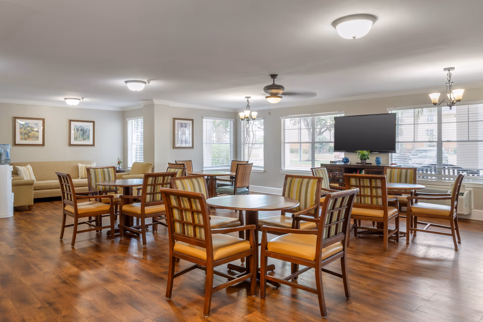 Bright communal dining/activity room with multiple round tables and wooden chairs, a seating area and wall-mounted TV.