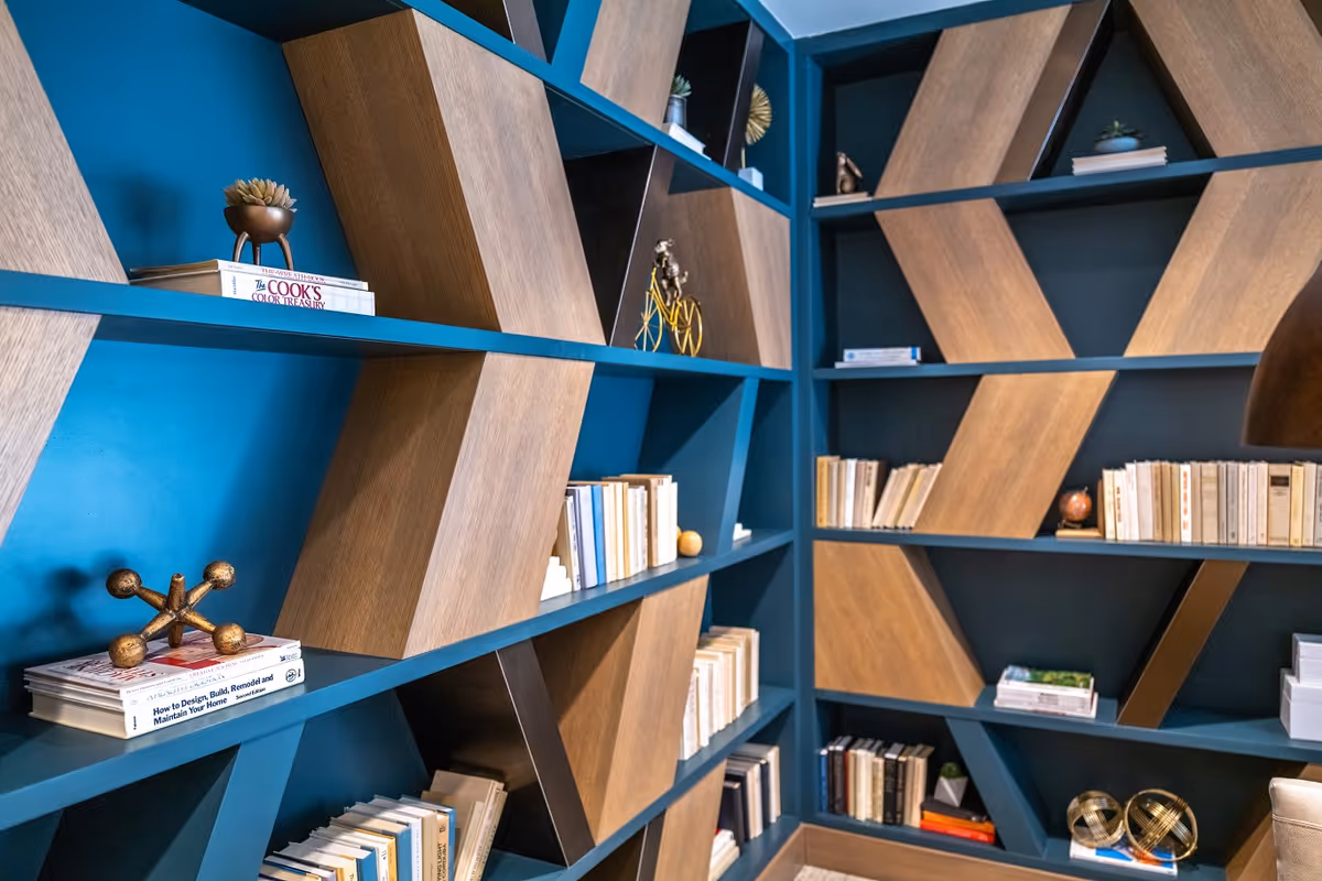 A modern bookshelf with a geometric design featuring blue shelves and wooden diagonal panels. The shelves hold various books, decorative objects including a small sculpture, a miniature bicycle, and small plants.