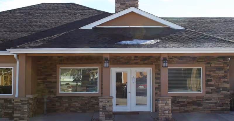Front exterior view of a single-story building with a stone facade, two large windows on either side of a set of double glass doors, a dark shingled roof, and two wall-mounted lantern-style lights flanking the entrance.