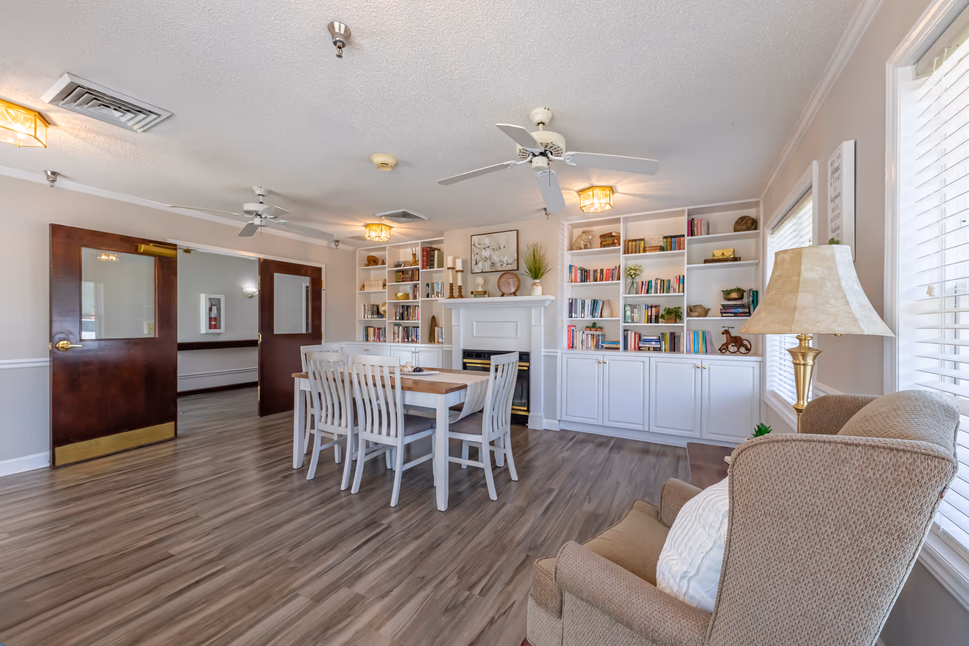 A bright and spacious common area with a wooden dining table and six white chairs in the center. The room features built-in white bookshelves filled with books and decorative items, a white fireplace, ceiling fans, and large windows with white blinds. There is a beige armchair with a white pillow and a gold lamp with a beige lampshade near the window. The floor is covered with wood-style vinyl planks, and double wooden doors with glass panels are visible on the left side.