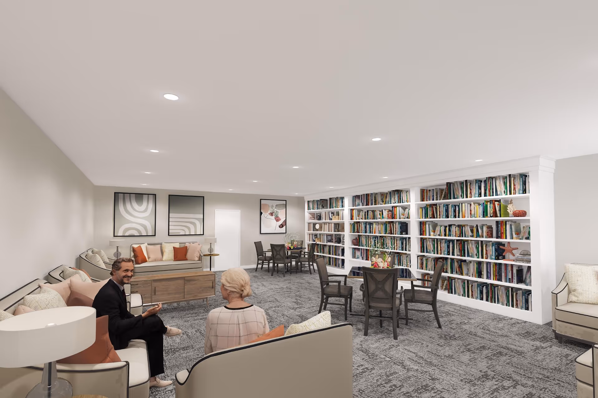 A spacious, well-lit common area with gray carpet and white walls. The room features several seating arrangements including sofas and chairs with cushions. There are multiple round tables with chairs around them. One wall is lined with built-in bookshelves filled with books and decorative items. Two people are sitting and conversing on a sofa in the foreground. The room has modern recessed lighting and framed artwork on the walls.