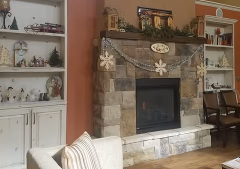 Cozy lounge area with a stone fireplace decorated with garland and snowflake ornaments, flanked by white display shelves and seating.