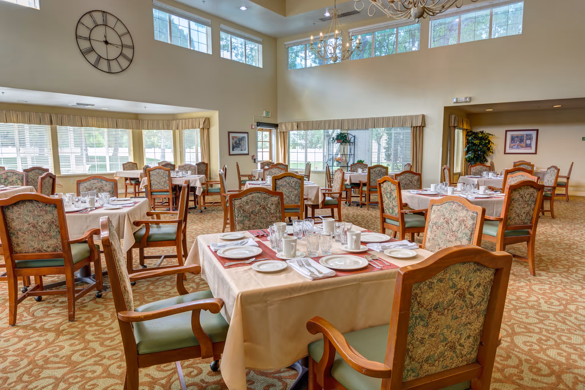 A bright and spacious dining room with multiple tables covered in beige tablecloths, each set with plates, glasses, cups, and silverware. The chairs have wooden frames with floral upholstered backs and green cushioned seats. Large windows with blinds and valances allow natural light to fill the room. A large clock is mounted on the wall, and a chandelier hangs from the ceiling.