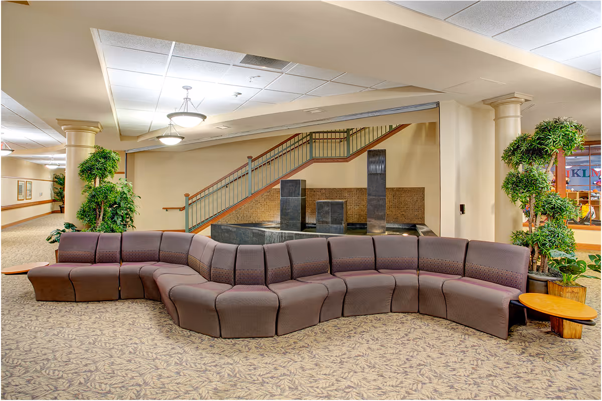 A spacious indoor common area with a curved sectional sofa in the center, beige walls, carpeted floor with a leaf pattern, potted plants on either side, and a staircase with a metal railing in the background. There are three modern pendant lights hanging from the ceiling and a water feature with three vertical stone pillars behind the sofa.