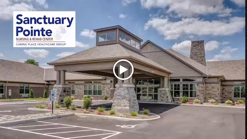 The front entrance of Sanctuary Pointe Nursing & Rehab Center with a covered porte-cochere, stone columns and parking area under a partly cloudy sky.