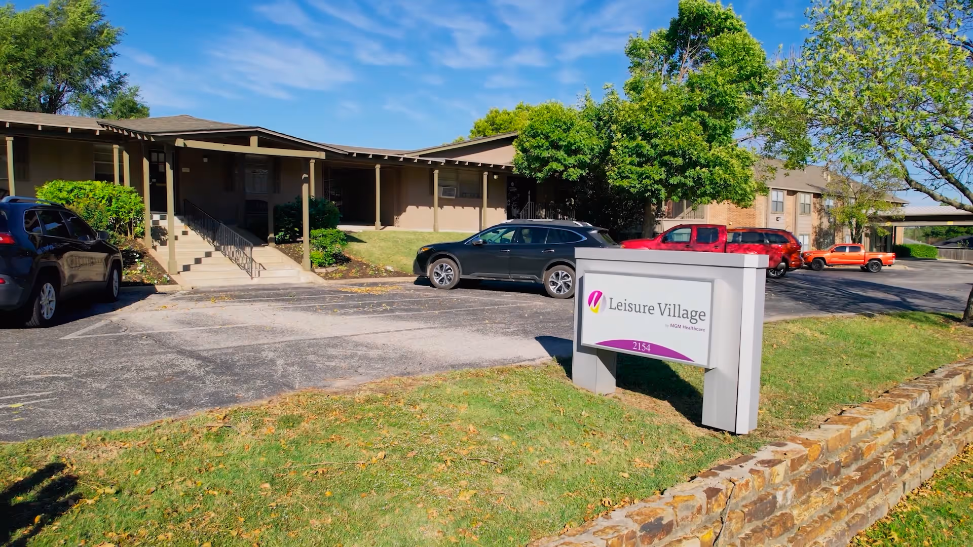 Front exterior of the Leisure Village health care building with a parking lot, several cars, lawn and a freestanding sign displaying the facility name.