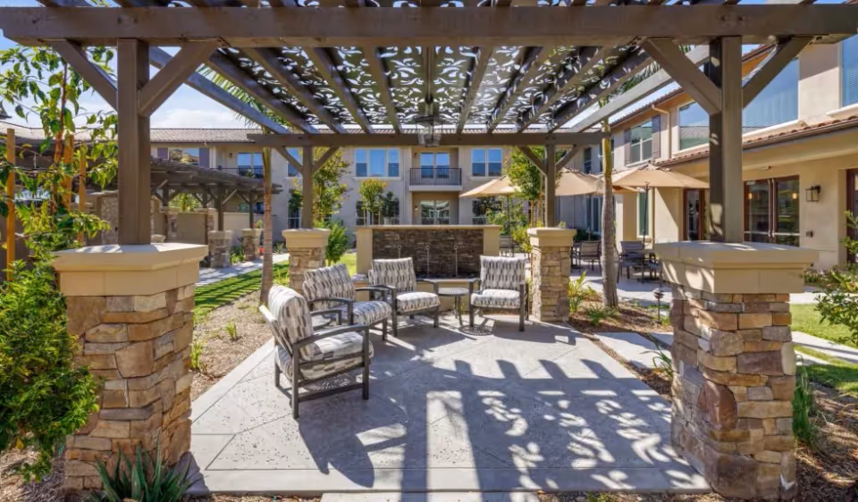 Outdoor courtyard with a pergola-shaded seating area of cushioned chairs and a water feature in front of the senior living building.