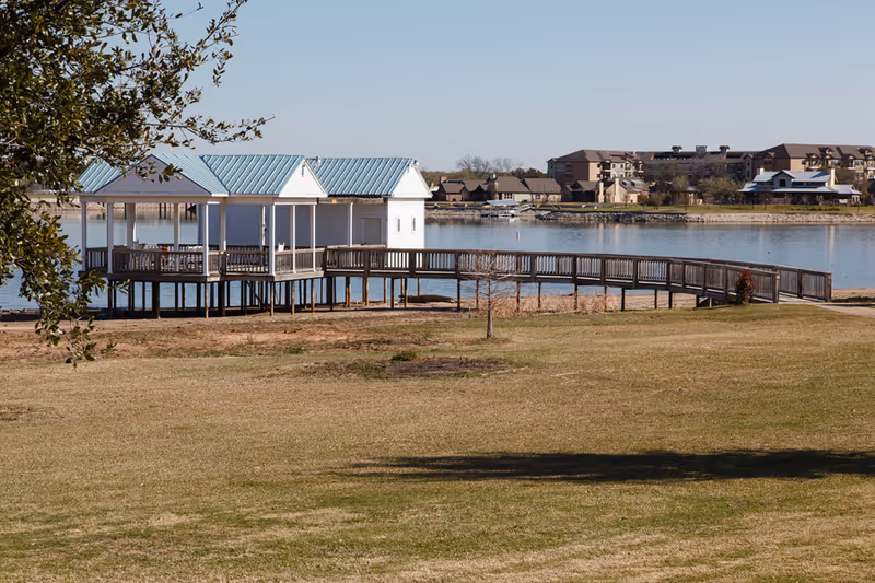 A wooden pier with a covered gazebo structure extending over a calm lake, surrounded by grassy land and trees, with residential buildings visible across the water under a clear blue sky.