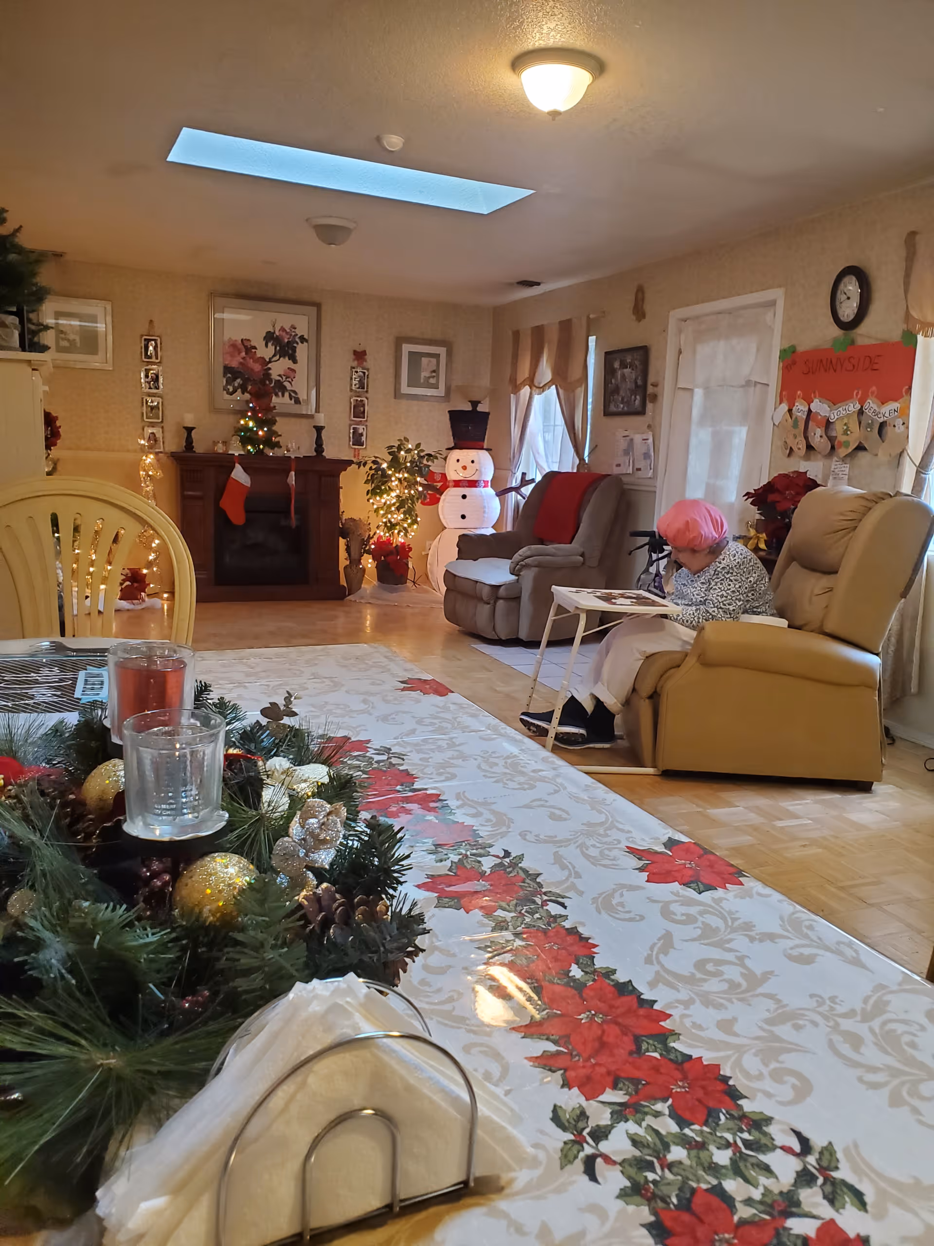 A cozy living room decorated for Christmas with a poinsettia-themed tablecloth on the dining table in the foreground. There is a festive centerpiece with candles and ornaments on the table. In the background, a woman with pink hair is seated in a beige recliner chair, reading or looking at something on a small table in front of her. The room features a fireplace with Christmas stockings hanging, a lit-up snowman decoration, and various framed pictures on the walls. The space is warmly lit with a ceiling light and natural light from windows with curtains.