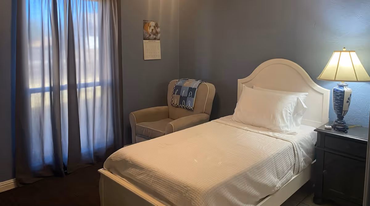 Small bedroom with a single bed, white headboard, bedside table and lamp, an armchair, and a curtained window.