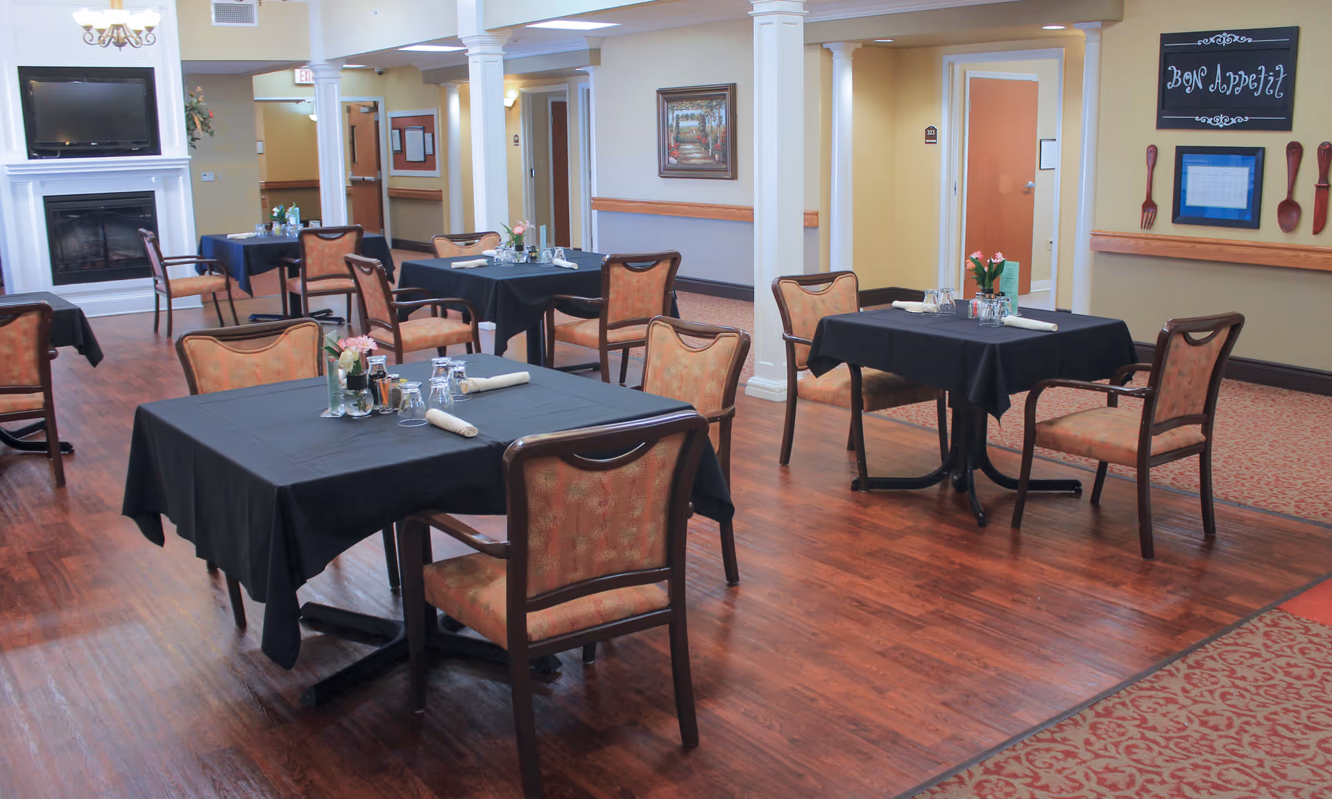 Dining room with multiple tables covered in black tablecloths and upholstered chairs on hardwood floors.