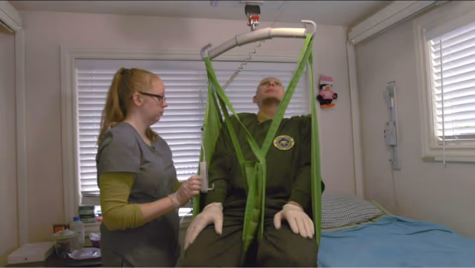 A caregiver assists a patient who is seated in a green sling lift inside a small room with a bed. The caregiver is adjusting the lift mechanism while the patient looks upward. The room has white walls, two windows with blinds, and a bed with blue bedding.