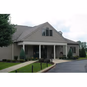 Front entrance of a single-story assisted living building with a covered porch, columns, landscaping, and a driveway.